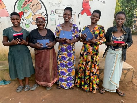 Five smiling women standing together outdoors, each holding a small gift box or item, wearing colorful traditional dresses