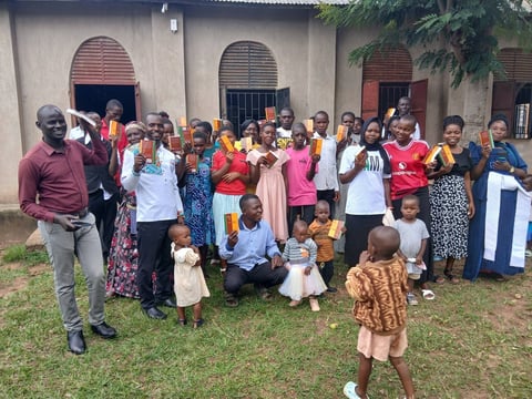 Large group of adults and children in colorful clothing standing together outside a white building with red-trimmed doors