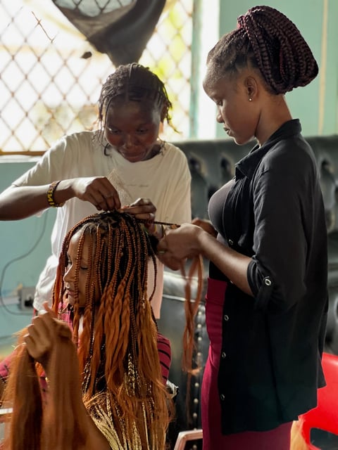 A woman in a black shirt braiding a childs reddish-brown hair indoors near a window with a mesh screen