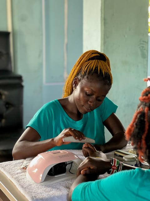 Woman in teal shirt filing or working on nails at a table with a pink nail lamp