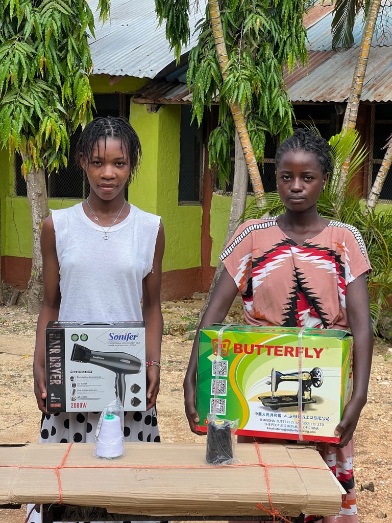 Two young women holding hair dryer and sewing machine boxes outdoors in front of a green and brown building